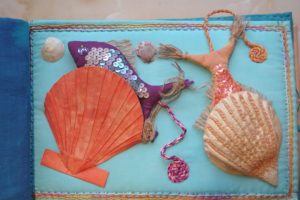 A colorful textile artwork featuring a sea-themed design. On a light blue fabric background, there are two large shell shapes. The left shell is orange with a pleated texture, and the right shell is beige with a textured surface. Between the shells, there is a purple fish adorned with shiny sequins, giving it a sparkling appearance. The fish has a small eye and a tail made of frayed fabric. There are also small, round, textured elements resembling sea urchins or coral. The edges of the fabric are bordered with multicolored stitching, adding a decorative frame to the piece.