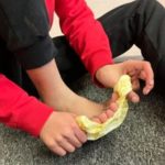 The image shows a person sitting on a carpeted floor with their legs bent. They are wearing a red long-sleeve shirt and black pants. The person is holding a piece of crumpled yellow tissue or paper towel with both hands, using it to wipe or clean their bare foot. The focus is on the hands and foot.