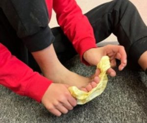 The image shows a person sitting on a carpeted floor with their legs bent. They are wearing a red long-sleeve shirt and black pants. The person is holding a piece of crumpled yellow tissue or paper towel with both hands, using it to wipe or clean their bare foot. The focus is on the hands and foot.