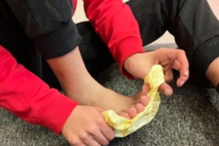 The image shows a person sitting on a carpeted floor with their legs bent. They are wearing a red long-sleeve shirt and black pants. The person is holding a piece of crumpled yellow tissue or paper towel with both hands, using it to wipe or clean their bare foot. The focus is on the hands and foot.