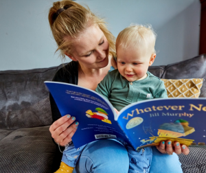 A mother sits with her young child reading a children’s book]
