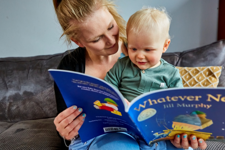 A mother sits with her young child reading a children’s book]